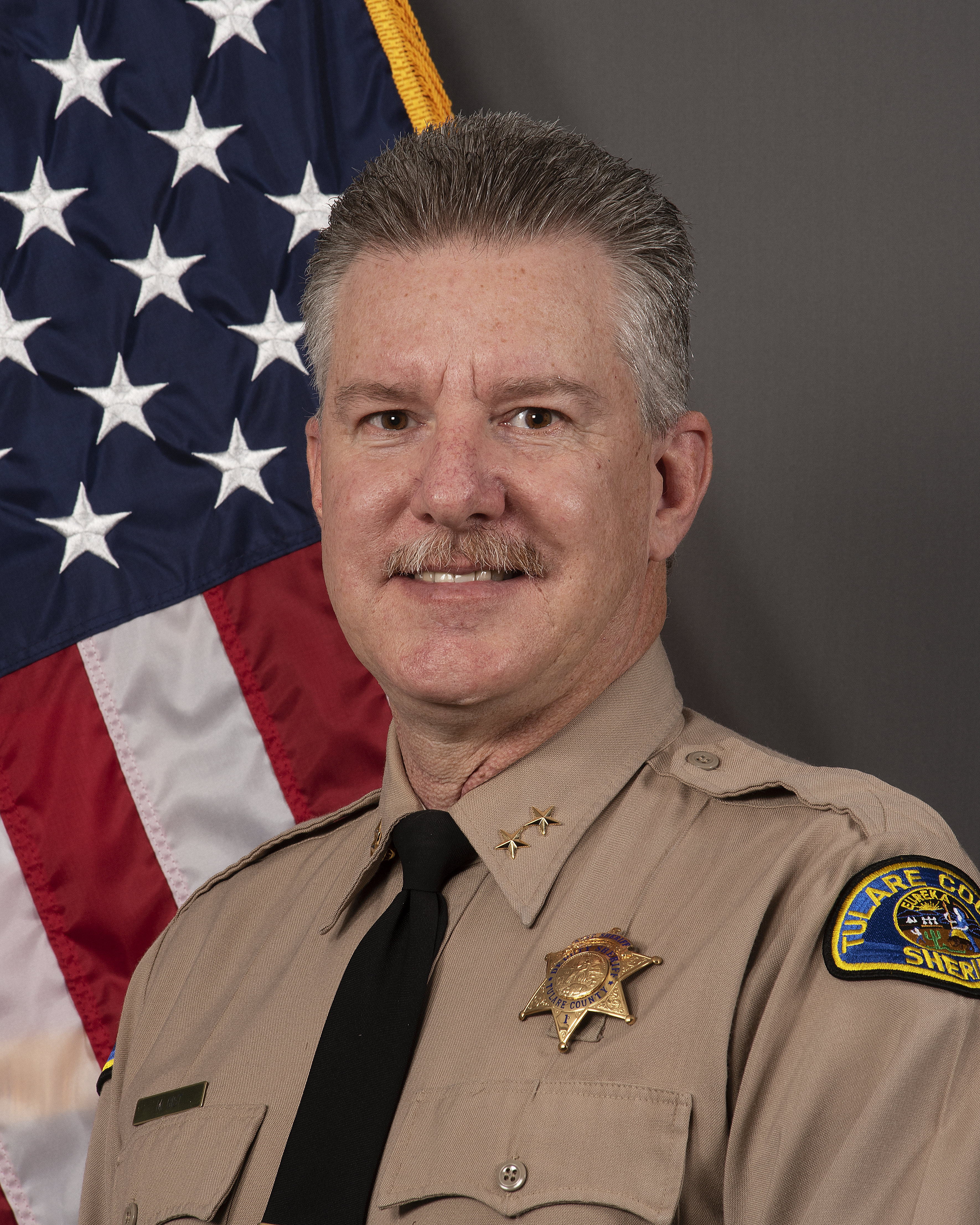 A head and shoulders portrait of Assistant Sheriff Mark Gist in uniform in front of an American Flag. 